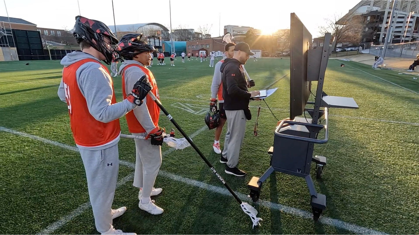 Two athletes in orange and gray uniforms with a coach on a sports field, using a video analysis system.